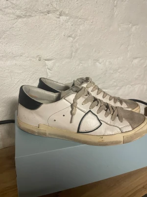 Philippe Model PRSX sneakers vit/svart - Säljer ett par Philippe Model PRSX Low Man sneakers i vitt och svart. Skorna har en rund tå, snörning och beige mockadetaljer på tungan och tåpartiet. Yttermaterialet är skinn med svarta detaljer vid hälen och en platt sula i gummi. Klassisk och stilren design.