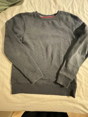 Mörkblå/grå sweatshirt från Tommy Hilfiger - Snygg mörkgråblå sweatshirt från Tommy Hilfiger med diskret logga broderad på bröstet. Tröjan har rund hals, ribbade muddar vid ärmslut och nederkant samt långärmad passform. Perfekt för dig som gillar stilrena och klassiska plagg. 