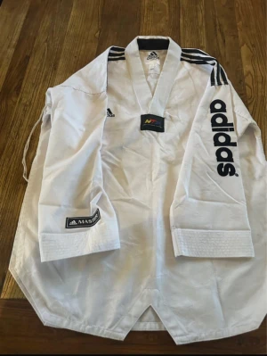 Vit Adidas taekwondodräkt 180cm - Säljer en vit taekwondoskjorta från Adidas med svarta detaljer och logga på bröstet och ärmen. Skjortan har klassisk v-ringad krage och svarta ränder på axlarna. Materialet är 70% bomull och 30% polyester, perfekt för träning. Inproderade teakwondologgor intyget. Använda två gånger nypris 3200:-