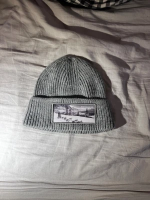Grå ribbad mössa från Revenite - Snygg grå ribbstickad mössa från Revenite med uppvikt kant och ett svartvitt tygmärke framtill som visar ett vintermotiv. Mössan är tillverkad i mjuk syntet och passar perfekt för kalla dagar. Enkel, stilren design som funkar till alla jackor.