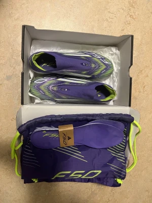 Adidas F50 lila fotbollsskor - Säljer ett par Adidas F50 fotbollsskor i lila med neongröna och vita detaljer. Skorna har en strömlinjeformad design, platt sula med dobbar och mönstrad ovandel. Kommer med matchande skopåse och extra innersulor. Perfekt för dig som vill sticka ut på planen.