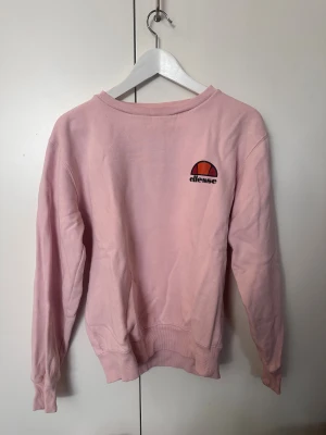 Ljusrosa sweatshirt från Ellesse - Säljer en ljusrosa sweatshirt från Ellesse med klassisk logga på bröstet. Tröjan har rund hals, långa ärmar och ribbade muddar vid ärmslut och nederkant. Perfekt för dig som gillar sportig och avslappnad stil.