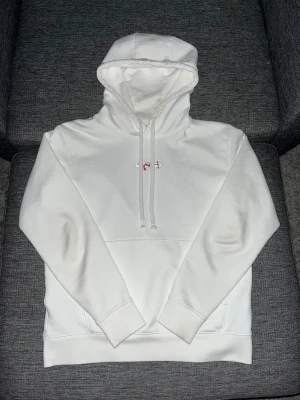 Vit hoodie från Hugo Boss - Vit hoodie från Hugo boss med röd broderad logga på bröstet och silverdetalj. Tröjan har snörning i huvan, magficka och ribbade muddar vid ärmslut och nederkant. Perfekt för dig som gillar cleana och stilrena plagg med en liten twist.