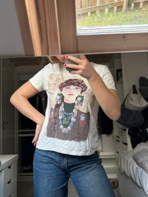 T-shirt med tryck - Vit t-shirt från Zara med ett konstnärligt tryck på framsidan som föreställer en person med detaljerade smycken och mönster. Rund hals och korta ärmar. Materialet är mjuk bomull och passformen är normal. Perfekt för dig som gillar unika prints!! Varan är använd men fortfarande bra skick 