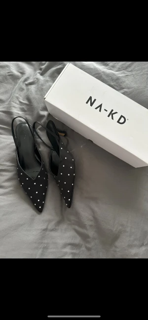 Svarta prickiga pumps från NA-KD - Snygga svarta pumps från NA-KD med spetsig tå och låg klack. Skorna har ett coolt prickigt mönster i vitt och en öppen häl med smal rem. Materialet är slätt och ger en elegant vibe, perfekt för dig som gillar att sticka ut med detaljer.