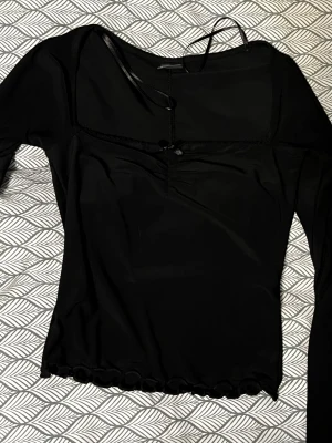 Svart långärmad topp med rosett Gina Tricot - Supersnygg svart långärmad topp från Gina Tricot i storlek XS. Toppen har en liten söt rosett framtill, volangkant nertill och smala band vid halsen. Materialet är mjukt och stretchigt, troligtvis polyester. Perfekt att styla med jeans eller kjol för en cool look.