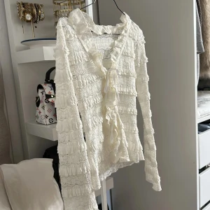 Vit spetsblus med volanger och knyt - Romantisk vit blus i spets med långa ärmar, volangdetaljer över hela och v-ringning med knyt framtill. Toppens material är genombruten spets och den har en feminin, omlottliknande look. Perfekt för dig som gillar en söt och trendig stil.