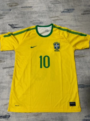 Brasilien Ronaldinho #10 Nike fotbollströja - Gul Brasilien fotbollströja från Nike med gröna detaljer och nummer 10 på både fram- och baksidan. RONALDINHO står tryckt på ryggen. Broderat CBF-emblem på bröstet, grön krage och axelband. Tillverkad i lätt Dri-Fit polyester för maximal komfort på planen.
