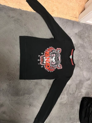 Svart Kenzo sweatshirt - Svart sweatshirt från Kenzo med ikoniskt broderat tigerhuvud i rött, rosa och vitt på bröstet samt texten 'KENZO PARIS'. Tröjan har rund hals, långa ärmar och ribbade muddar. Materialet är mjuk bomull och insidan har en röd detalj vid halsen.