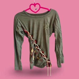 Grön långärmad tröja med snörning - Unik grön långärmad tröja med snörning i kontrastfärg längs sidan och broderade detaljer med blad och mönster. Tröjan har en rund hals och cool asymmetrisk design som sticker ut. Perfekt för dig som gillar boho-vibe och detaljer.