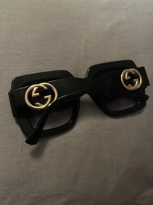 Svarta fyrkantiga solglasögon från Gucci - Säljer ett par svarta, Gucci solglasögon med fyrkantig form och stora guldiga GG-loggor på sidorna. Glasögonen har mörka linser och en riktigt statement look. Modellnummer GG1023SA. Perfekta för dig som vill sticka ut. Köptes för 3800 säljs för 1300 (fått som pressent) , pris kan diskuteras vid snabb affär. Kvitto finns tyvärr ej men man kan söka på serienumret. Först till kvarn, går lägst ner till 1000 vid snabb affär