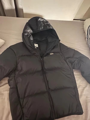 Svart pufferjacka från Lacoste - Svart pufferjacka från Lacoste med huva och dragkedja. Jackan har det klassiska Lacoste-krokodilmärket på bröstet och en stor broderad krokodil på ryggen. Fylld med syntetmaterial för extra värme, perfekt för kalla dagar. Stilren och enkel design.