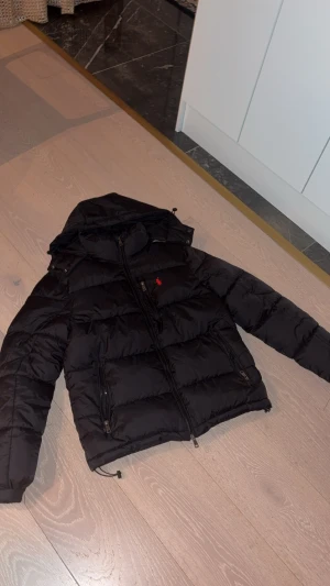 Svart pufferjacka från Polo Ralph Lauren - Svart pufferjacka från Polo Ralph Lauren med huva och dragkedja framtill. Jackan har det klassiska röda Polo-logot på bröstet och flera praktiska fickor med dragkedja. Perfekt för kalla dagar och har en snygg, sportig look. Storlek S.