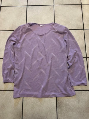 Ljuslila långärmad tröja med mönster storlek M/L - Ljuslila långärmad tröja med diskret geometriskt mönster i samma ton. Tröjan har en rundad halsringning och är tillverkad i ett mjukt material som känns skönt mot huden. Perfekt för dig som gillar en chill och enkel stil med lite extra detaljer.