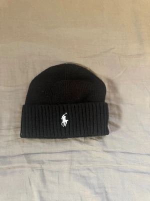 Svart stickad mössa från Polo Ralph Lauren - Svart ribbstickad mössa från Polo Ralph Lauren med klassisk vit broderad logga framtill. Mjuk och varm, perfekt för kyliga dagar. Enkel design med uppvikt kant som ger en clean och stilren look.