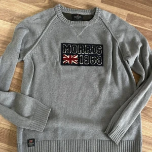 Grå bomullströja Morris 1968 - Grå långärmad tröja från Morris med broderad patch framtill där det står 'Morris 1968' och en brittisk flagga. Tröjan är i 100% bomull, har ribbade muddar och en klassisk rund hals. Perfekt för dig som gillar stilrena och sportiga plagg.