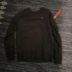 Svart sweatshirt från Alpha Industries - Svart sweatshirt från Alpha Industries med klassisk rund hals och lång ärm. Tröjan har en liten logotyp på bröstet och en ikonisk röd tag på ärmen. Perfekt för dig som gillar streetwear och enkel stil.