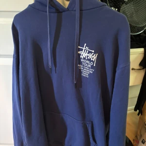 Blå Stüssy hoodie med tryck - Säljer en blå hoodie från Stüssy med vit logga och text på både bröstet och ryggen. Tröjan har huva med snörning och en stor magficka. Perfekt för dig som gillar streetwear och vill sticka ut med en ikonisk look.
