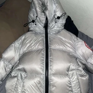 Silver dunjacka från Canada Goose - Säljer en riktigt fet silverfärgad dunjacka från Canada Goose. Jackan har stor huva med dragsko, svart dragkedja och klassisk patch på ärmen. Perfekt för kalla dagar och har en snygg, puffig look som sticker ut. Insidan är svart och jackan har reflekterande detalj på huvan. Finns kvitto om du vill ha den 
