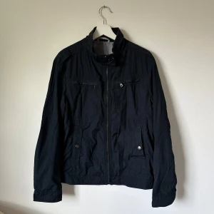 Mörkblå vindjacka från Hugo Boss - Stilren mörkblå vindjacka från Hugo Boss med hög krage, dragkedja framtill och flera fickor med knappdetaljer. Jackan har ett lätt och smidigt material, perfekt för övergångssäsonger. Klassisk design med diskret logga på insidan. 2I7