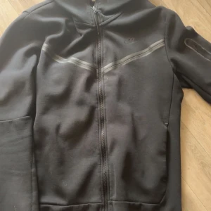 Svart Nike hoodie med dragkedja - Svart hoodie från Nike med hel dragkedja framtill och huva. Snygga diskreta detaljer över bröstet och på ärmen. Perfekt för dig som gillar sportig och clean stil. Mjuk och bekväm, passar till allt.