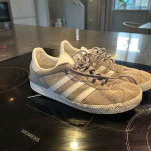 Adidas Gazelle grå/vita sneakers mocka - Säljer ett par Adidas Gazelle sneakers i grå mocka med vita detaljer och klassiska tre ränder på sidorna. Skorna har vit plös och hälparti med Adidas-logga, samt snörning framtill. Perfekta för dig som gillar retrostil och vill ha något stilrent till vardags.