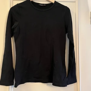 Svart långärmad tröja från Lindex - En stilren svart långärmad tröja från Lindex i mjuk bomull. Tröjan har rund halsringning och en klassisk passform som funkar till allt. Perfekt basplagg att ha i garderoben för alla tillfällen.