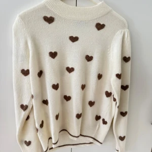 Vit stickad tröja med bruna hjärtan - Supersöt vit stickad tröja med bruna hjärtan över hela. Tröjan har rund halsringning, långa ärmar och ribbade muddar. Perfekt för dig som gillar gulliga detaljer och vill ha en mysig look.
