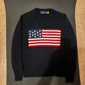 Mörkblå stickad tröja Polo Ralph Lauren - Mörkblå stickad tröja från Polo Ralph Lauren med amerikansk flagga i rött, vitt och blått på bröstet samt RL-broderi. Tröjan är i 100% bomull, har rund hals och långa ärmar med ribbade muddar. Perfekt för dig som vill sticka ut med en klassisk preppy stil.