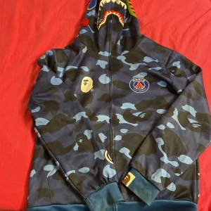 PSG x Bape blå camo hoodie - Säljer en blå camo hoodie DOPE, Tröjan har dragkedja, ribbade muddar och är tillverkad mjuk material. Har använt den ett par gånger använder inte längre. Köpte för 500kr. Pris kan diskuteras.