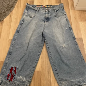 JNCO wide jeans med brodyr, 26" - Säljer ett par ikoniska JNCO jeans i ljusblå denim med bred passform och stora bakfickor. Byxorna har slitna detaljer, broderad logga i rött och svart på benet samt coolt mönstrat innerband i midjan. Perfekta för dig som gillar streetwear och 90-talsstil.