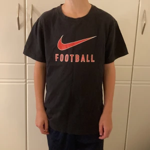 Svart Nike t-shirt med röd logga - Svart t-shirt från Nike i bomull med röd Nike Swoosh och texten 'FOOTBALL' på bröstet. Klassisk passform med rund hals och korta ärmar. Säljer pågrund av att den inte kommer/kommit till användning.