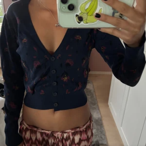 Mörkblå blommig kofta från Zara - Säljer en mörkblå croppad kofta från Zara med blommigt mönster i rött och lila. Koftan har djup v-ringning, långa ärmar och knappar framtill. Perfekt för lager-på-lager och snygg till både jeans och kjol.