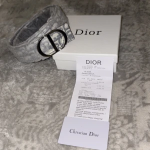 Grått Dior bälte med monogram - Snyggt bälte från Christian Dior i grått och vitt med ikoniskt Dior-monogram och silverfärgat D-spänne. Bältet har en exklusiv känsla med läderdetaljer på insidan och passar perfekt för att lyfta din outfit med en lyxig touch.