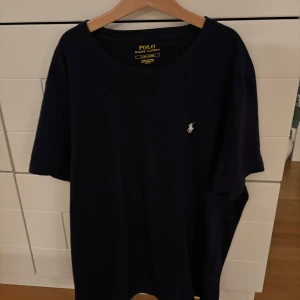 Mörkblå t-shirt från Polo Ralph Lauren - Snygg mörkblå t-shirt från Polo Ralph Lauren med klassisk vit logobrodyr på bröstet. Modellen har rund hals och korta ärmar, tillverkad i mjuk bomull för skön passform. Perfekt för dig som gillar stilrena och tidlösa plagg.