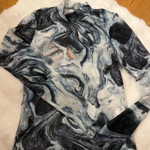 Marmor-mönstrad mesh polotröja new Yorker  - Säljer en tunn och stretchig mesh polotröja från new Yorker i storlek M. Tröjan har ett unikt marmor-inspirerat mönster i svart, grått, vitt och beige med glittriga detaljer. Långärmad och med hög krage, perfekt för att sticka ut i mängden.