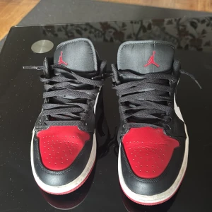 Nike Air Jordan 1 Low svart/röd/vit - Säljer ett par Nike Air Jordan 1 Low i klassisk svart, röd och vit färg. Skorna har svart snörning, röd tåbox och häl, vit sidopanel samt svart swoosh. Tillverkade i skinn med perforerad tå och ikonisk Jumpman-logga på plösen. Perfekta för dig som gillar streetwear.