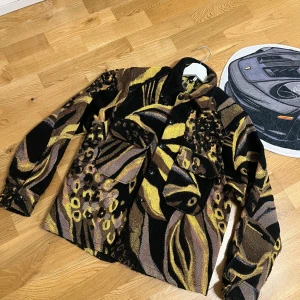 Animalmönstrad overshirt från Sunflower - Overshirt från Sunflower med djurinspirerat mönster i svart, gul och brun. Skjortan har stora bröstfickor, knappar framtill och är tillverkad i en mix av syntet, polyester, ull och polyamid. Perfekt statement-plagg med loose fit och lång ärm.