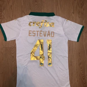 Palmeiras fotbollströja Puma S - Snygg vit Palmeiras fotbollströja från Puma med gröna detaljer och guldiga tryck. Tröjan har klubbmärke på bröstet, sponsortryck och nummer 41 med namnet Estêvão på ryggen. Tillverkad i lätt och ventilerande material, perfekt för match eller träning.