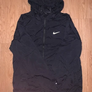 Svart Nike vindjacka med huva - Svart vindjacka från Nike med klassisk vit swoosh-logga på bröstet. Jackan har huva med dragsko, hel dragkedja framtill och är tillverkad i lätt polyester. Perfekt för blåsiga dagar och sportiga outfits.