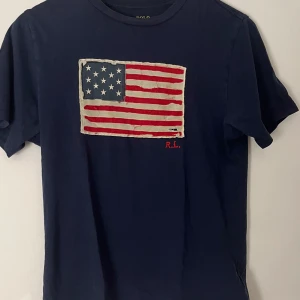 Mörkblå t-shirt Polo Ralph Lauren - Mörkblå t-shirt från Polo Ralph Lauren med broderad amerikansk flagga framtill och RL-detalj i rött. Klassisk rund hals och korta ärmar. Tillverkad i mjuk bomull för en skön känsla. Bara att höra av sig vid frågor, priset är absolut inte hugget i sten