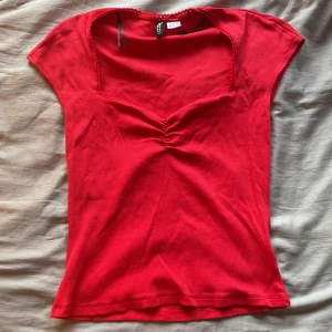 Röd topp med v-ringning från H&M - Snygg röd topp från H&M med v-ringning och rynkdetalj framtill. Toppen har korta ärmar och dekorativ virkad kant runt halsen. Tillverkad i mjuk bomull, perfekt för dig som gillar färg och enkel stil. Köpt för 99kr