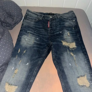 Dsquared2 slitna blå jeans - Riktigt snygga blå jeans från Dsquared2 med slitna partier och coola färgstänk. Klassisk femficksmodell med tydliga slitningar och en röd Dsquared2-tag vid gylfen. Jeansen har en rak passform och är tillverkade i jeansmaterial. har haft på mig dom en gång men var för stora