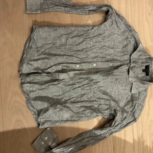 Grå linneskjorta  - Snygg grå skjorta i linne med klassisk krage och vita knappar framtill. Skjortan har lång ärm med manschett och en avslappnad passform. Perfekt för en chill och stilren look.