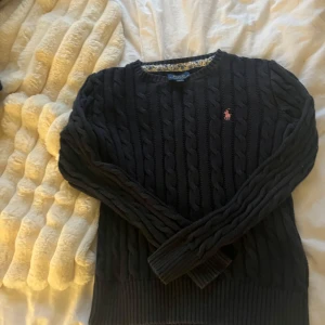 Svart kabelstickad tröja Polo Ralph Lauren - Kabelstickad svart tröja från Polo Ralph Lauren med klassisk rund halsringning och broderad rosa logga på bröstet. Tröjan har långa ärmar och ribbade muddar för en snygg passform. Perfekt för lager-på-lager under kyliga dagar.