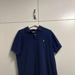 Marinblå piké från Ralph Lauren - Snygg marinblå pikétröja från Polo Ralph Lauren i custom fit. Klassisk krage, korta ärmar och gul broderad logga på bröstet. 