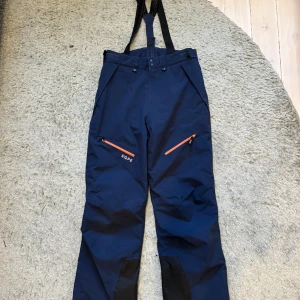 Mörkblå skidbyxor från EQPE - Mörkblå skidbyxor från EQPE med svarta förstärkningar vid bensluten och orange dragkedjor på fickorna. Byxorna har hängslen och är perfekta för vinterns äventyr. Funktionell design med justerbar midja och praktiska detaljer. Storlek 164/s.           Nypris 2000 