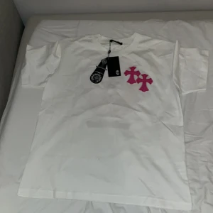 Vit Chrome Hearts t-shirt med rosa kors - Vit t-shirt från Chrome Hearts i bomull med två rosa kors på bröstet och stort rosa kors med logga på ryggen. Klassisk passform och korta ärmar. Snygga detaljer och tydlig streetwear-vibe.