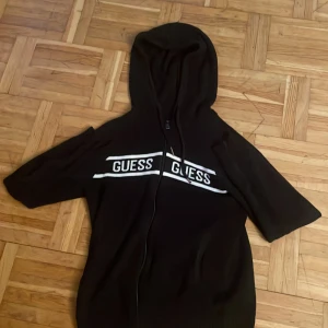 Svart hoodie från Guess med dragkedja - Svart hoodie från Guess i storlek XS med vit logga på bröstet. Tröjan har huva med snörning och hel dragkedja framtill. Tillverkad i mjukt stickat material, perfekt för en chill och avslappnad stil. Ordinare pris 1500.