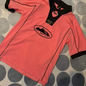 Rosa fotbollströja med svart krage - Säljer en rosa fotbollströja med svart krage och svarta detaljer. Tröjan har korta ärmar och ett tryckt motiv på bröstet. Tillverkad i ett lätt och sportigt material som passar perfekt för träning eller match.